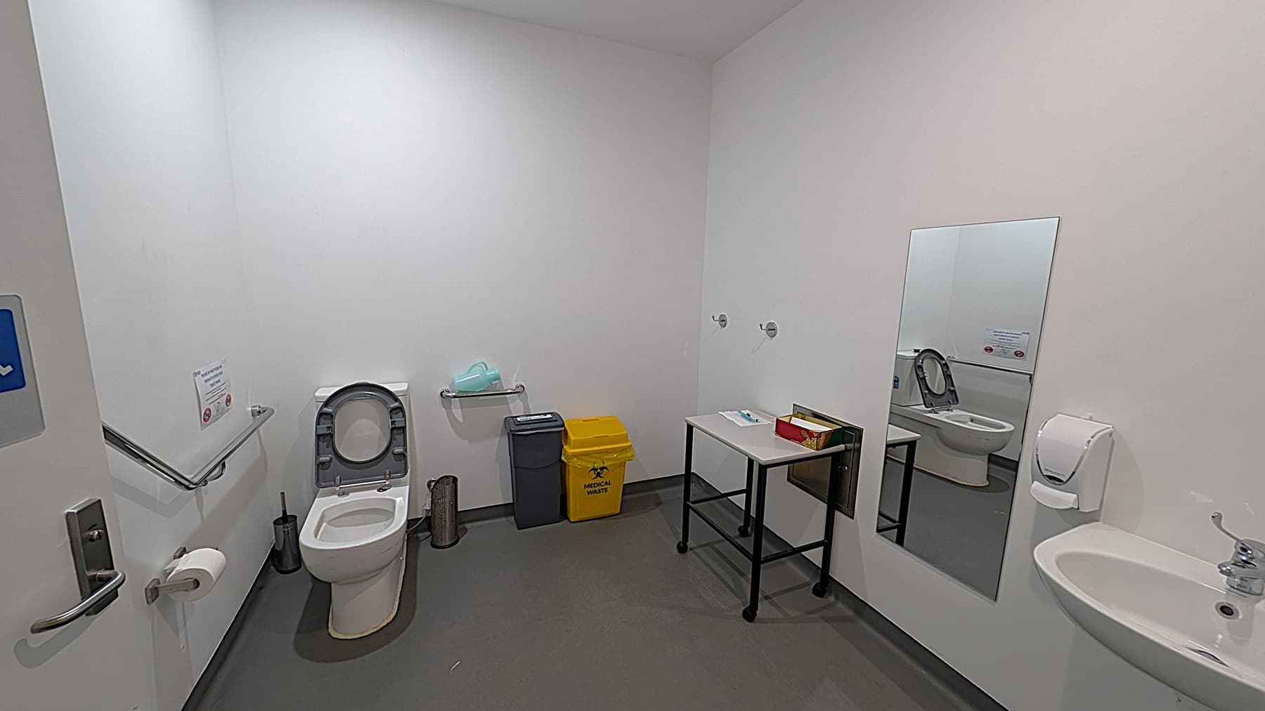 A clean, accessible bathroom with a white toilet on the left, equipped with a grab rail and toilet paper holder. Behind the toilet are a black bin and a yellow medical waste bin. A small table with a box of tissues is on the right. A large mirror is mounted on the wall, reflecting the toilet. A white sink with a soap dispenser is on the far right. The floor is grey, and the walls are white.