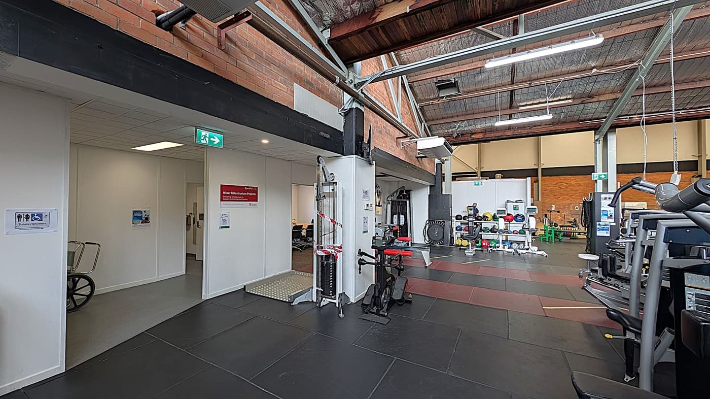 A gym interior with a high, industrial-style ceiling. On the left, there's a doorway leading to a hallway with a wheelchair sign and an exit sign above. A piece of gym equipment is partially visible near the entrance. In the centre, various gym machines and weights are arranged, including a cable machine with red and white tape. On the right, more gym equipment is visible, including a treadmill. The floor is covered with black and brown rubber mats, and the walls are brick.