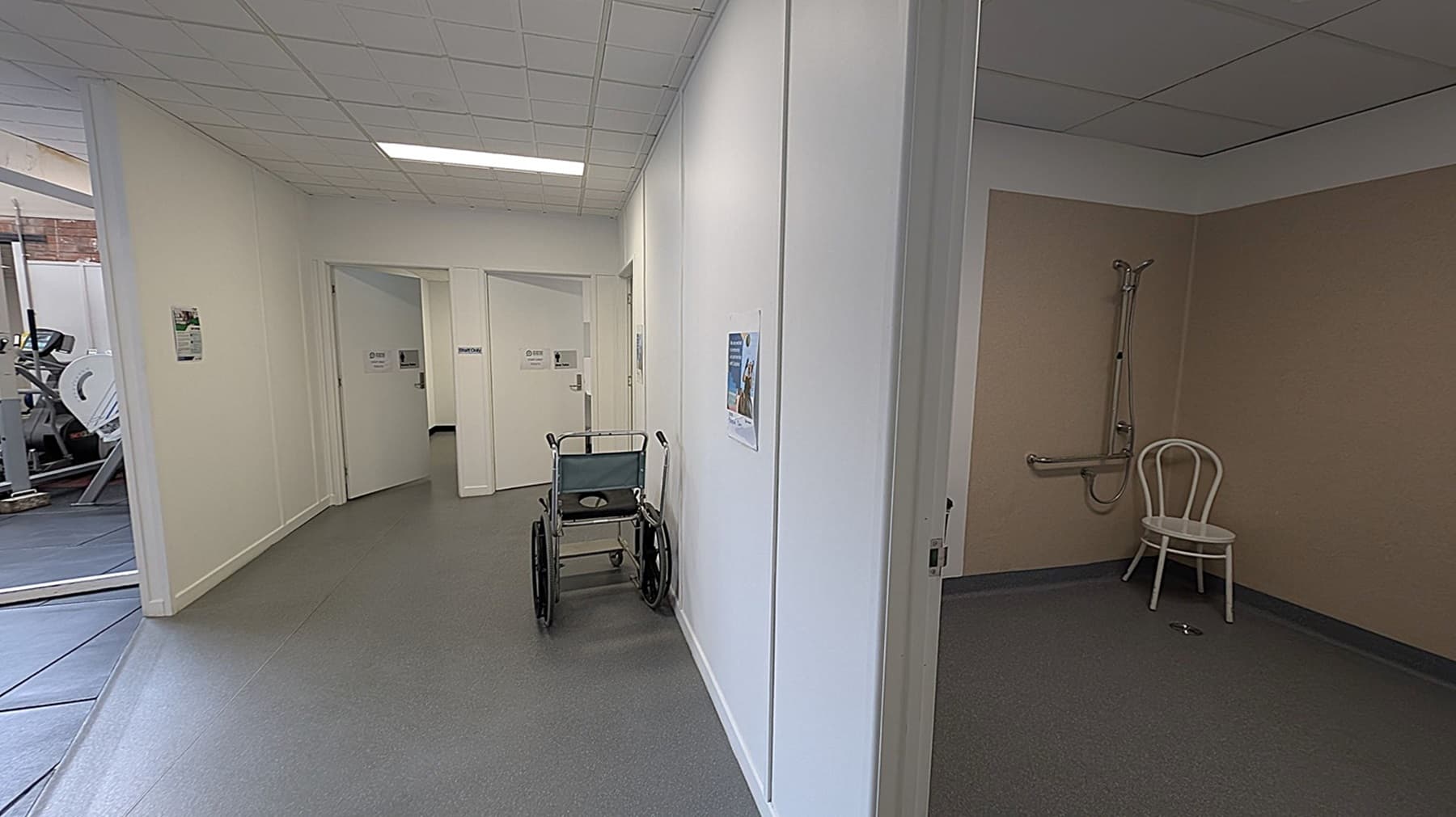 A hallway with grey flooring and a white ceiling. On the left, there are doors with signs and a glimpse of gym equipment through a glass partition. In the centre, a wheelchair is parked against the wall. On the right, an open room with beige walls features a showerhead and a white chair.