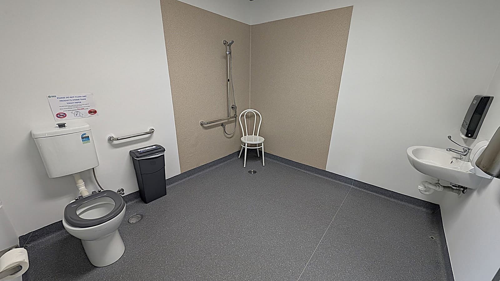 A public restroom with grey flooring and white walls. On the left, there's a toilet with a black seat, a grab bar, and a small bin. A sign above the toilet advises against flushing anything other than toilet paper. In the centre, a white chair is placed against the wall with a vertical grab bar. On the right, a small white sink with a lever tap is mounted on the wall, accompanied by a soap dispenser.