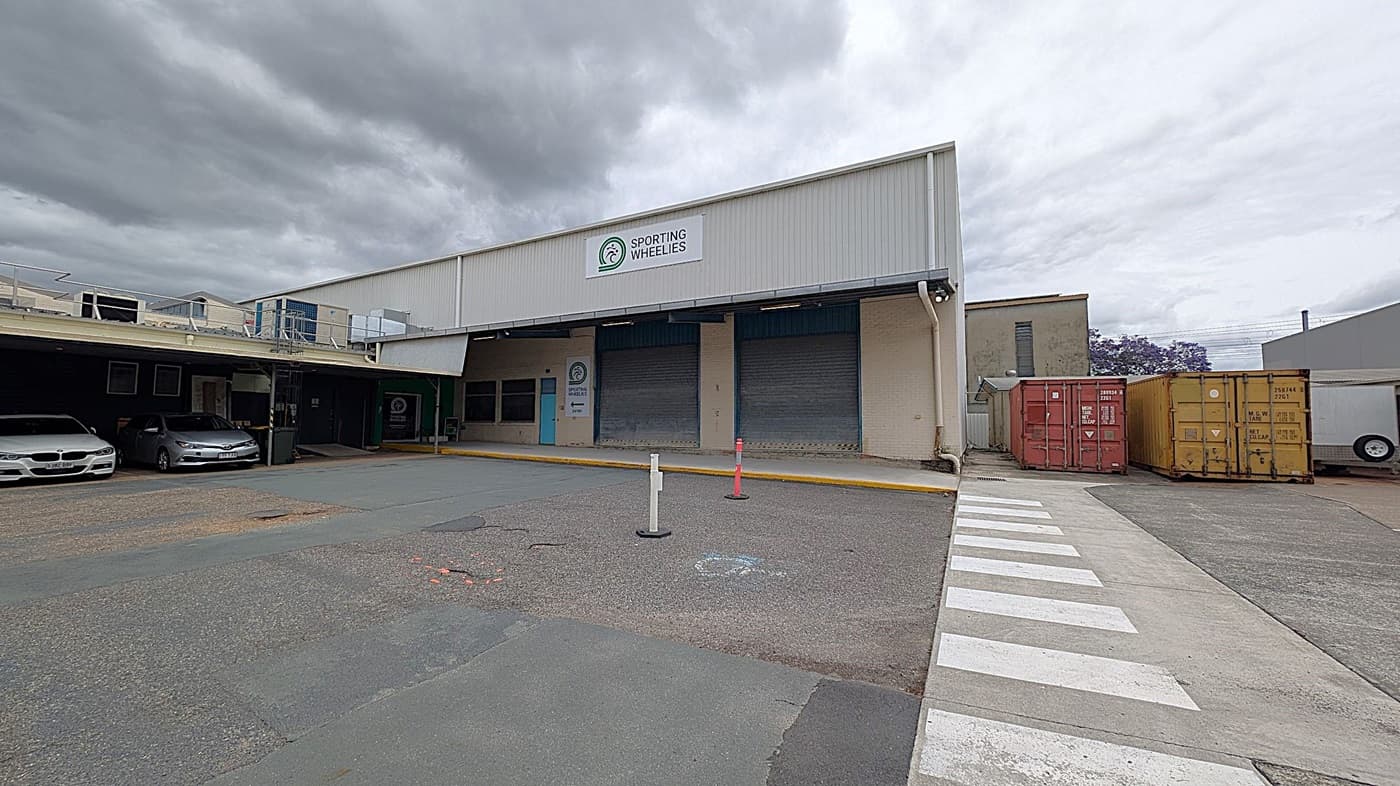 A warehouse with a sign reading "Sporting Wheelies" is in the centre. On the left, two cars are parked under a covered area. On the right, there are two shipping containers, one red and one yellow. A zebra crossing leads to the entrance, and there are two bollards in front of the building. The ground is paved, and the building has a corrugated metal exterior.