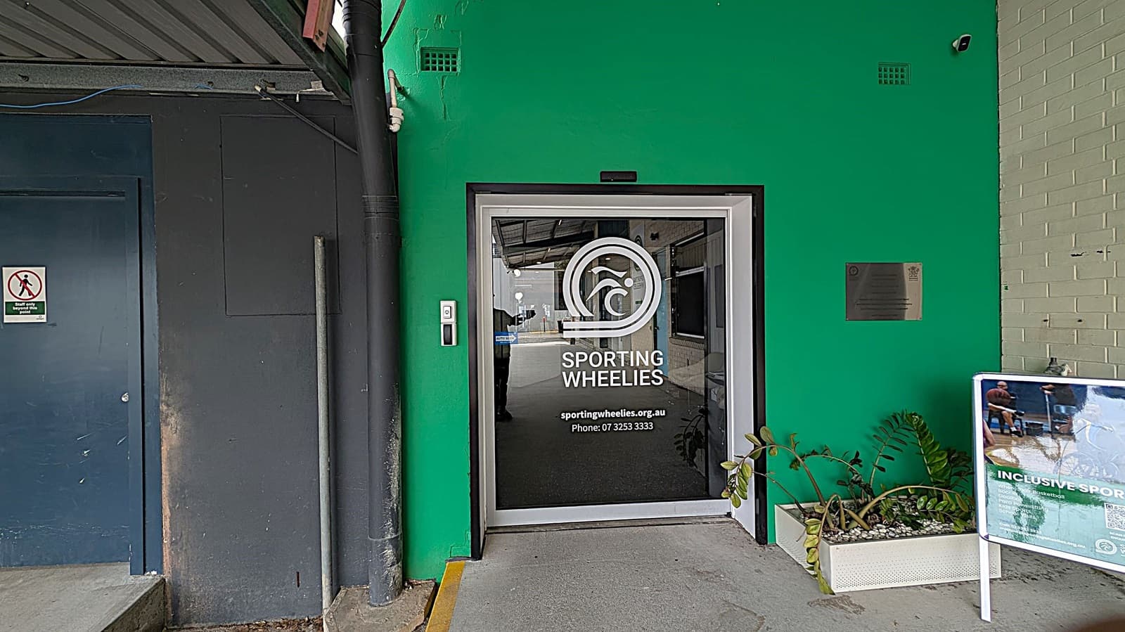 A green wall with a glass door in the centre, featuring the Sporting Wheelies logo and contact information. To the right, a plaque is mounted on the wall above a planter with green foliage. A signboard with images and text about inclusive sports is also on the right. On the left, a dark blue door with a "start your workout" sign is visible. The ceiling is industrial with exposed beams and pipes.