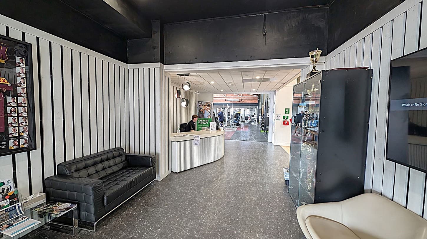 A reception area with a black leather sofa on the left, next to a wall with vertical white panels. A glass table holds magazines. In the centre, a curved reception desk with a person seated behind it, under three hanging lights. A poster about exercise therapy is visible. On the right, a glass cabinet displays trophies, with a large trophy on top. A beige chair is nearby. The floor is dark with a speckled pattern, and the ceiling is white with recessed lighting. In the background,