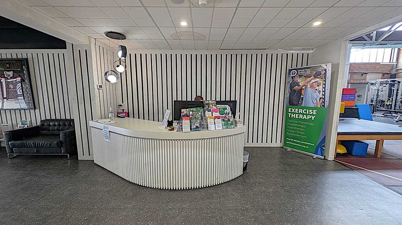 A reception area with a curved white desk in the centre, displaying brochures and a computer monitor. Behind the desk is a wall with vertical white slats. To the left, a black leather sofa and a framed sports jersey are visible. On the right, a green banner advertising exercise therapy stands near gym equipment. The floor is dark with a speckled pattern, and the ceiling is white with recessed lighting.