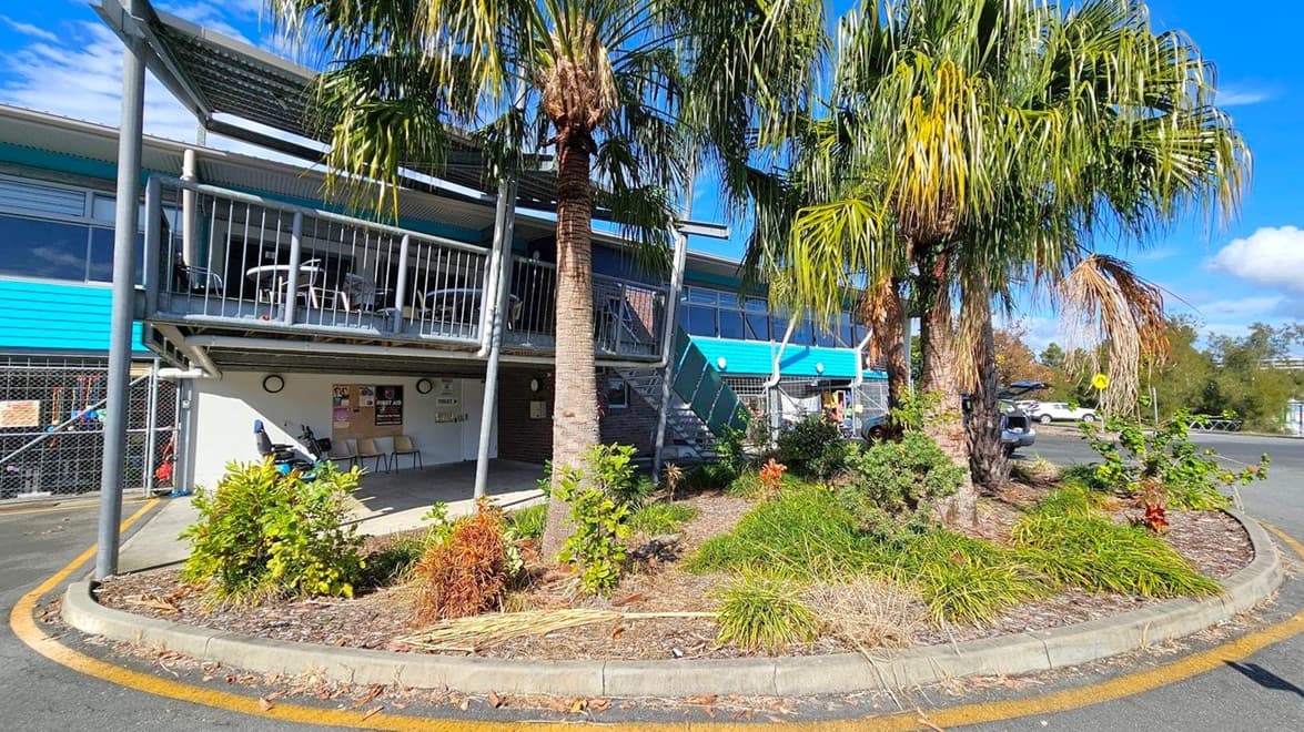 A building with a blue and grey exterior, featuring a balcony with metal railings and outdoor seating on the left. Below the balcony, there are posters and a first aid sign on the wall. In the foreground, a landscaped area with palm trees and various shrubs is surrounded by a kerb. To the right, a car park with a few vehicles is visible, along with a road and more greenery in the background.