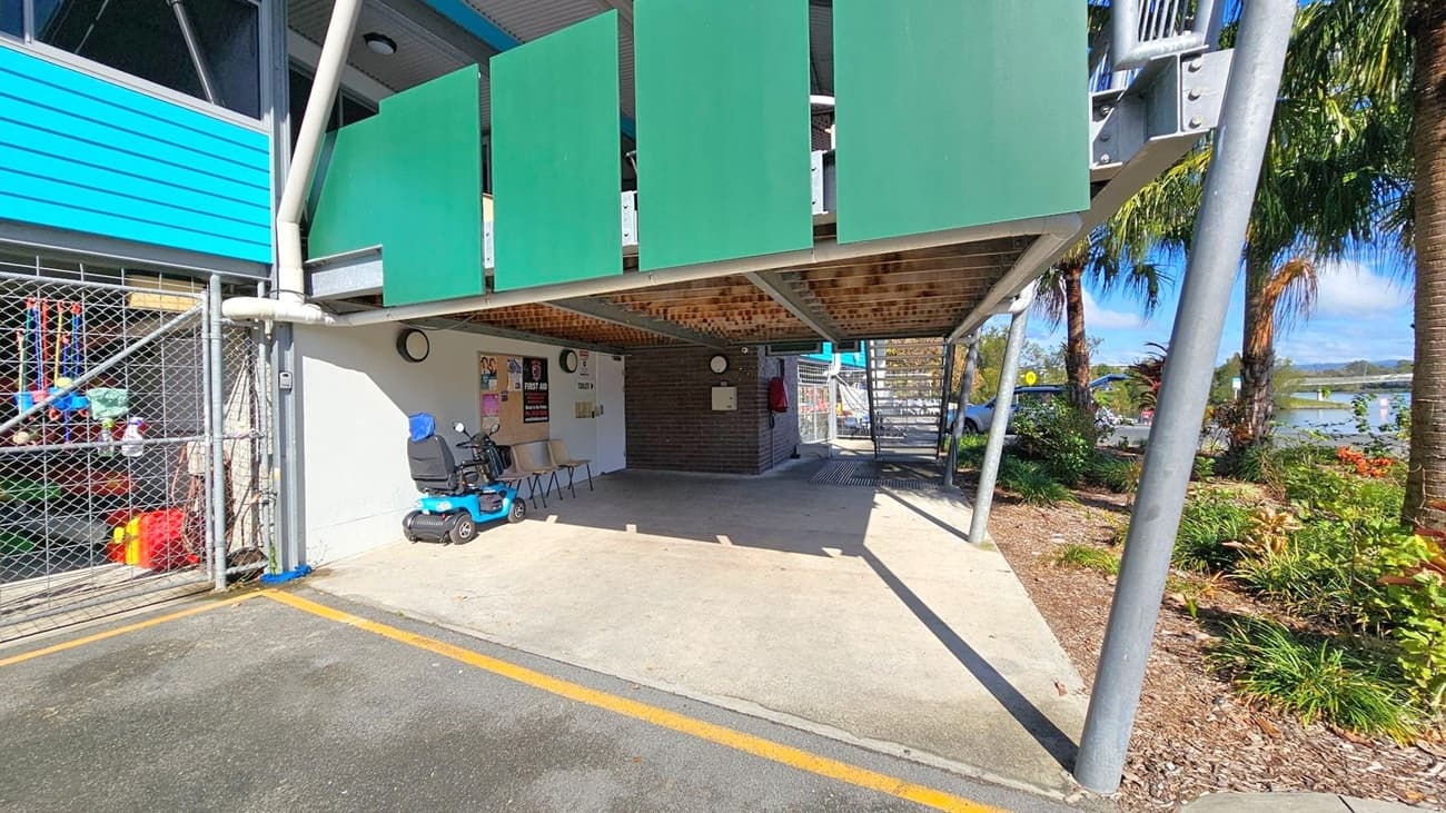 Under a building with green panels, there's a sheltered area with a blue mobility scooter and two chairs. A chain-link fence on the left encloses various items. The ground is a mix of concrete and asphalt with a yellow line. On the right, a garden with palm trees and shrubs is visible. A staircase leads up from the back, and a body of water can be seen in the distance. The ceiling is supported by metal beams.