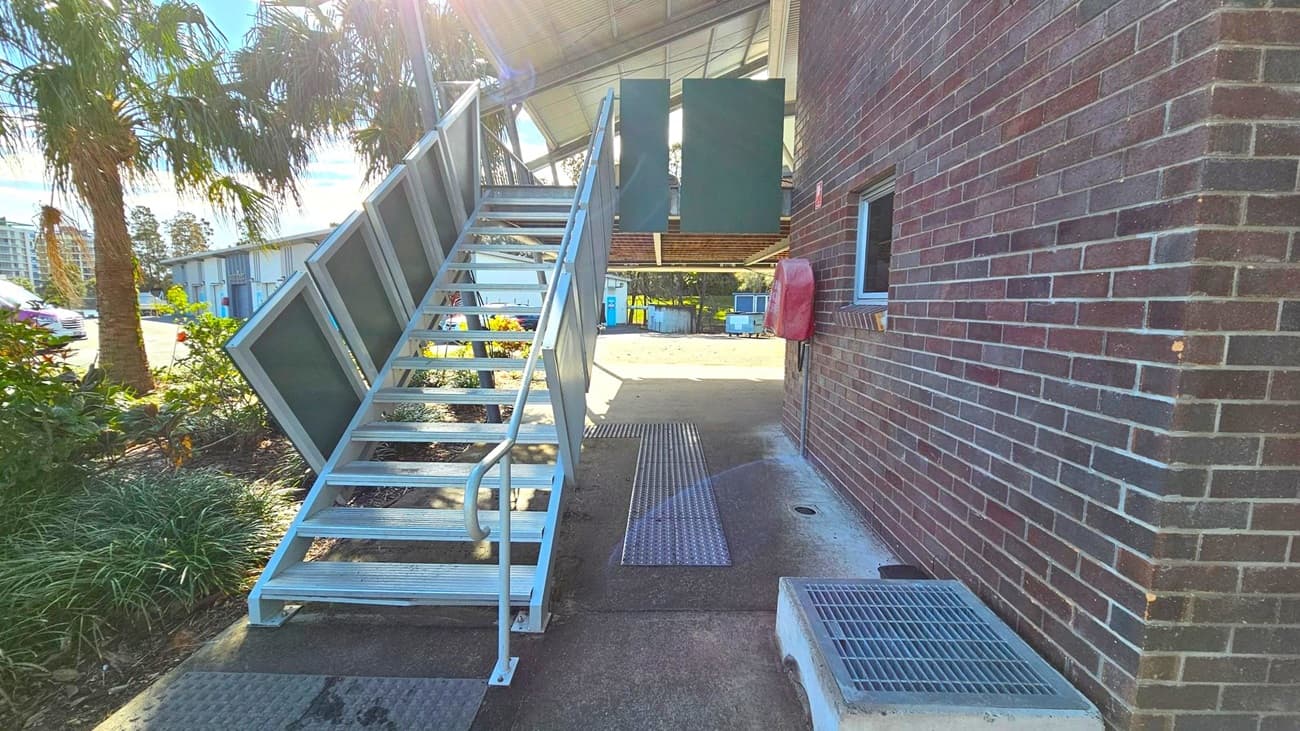 A metal staircase with a handrail on the left leads up to a building. The building has a brick exterior with a small window and a red box mounted on the wall. To the left of the stairs, there are palm trees and a garden area. In the background, there are industrial buildings and a parked car. The ground is concrete with metal grates, and the ceiling of the structure above is metal.