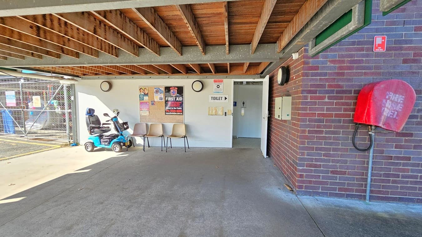 A covered area with a concrete floor and a wooden beam ceiling. On the left, a blue mobility scooter is parked next to three beige chairs. Posters and a clock are mounted on the white wall. A sign points to a toilet through an open door. On the right, a red fire hose reel is attached to a brick wall. In the background, a chain-link fence with various signs is visible.