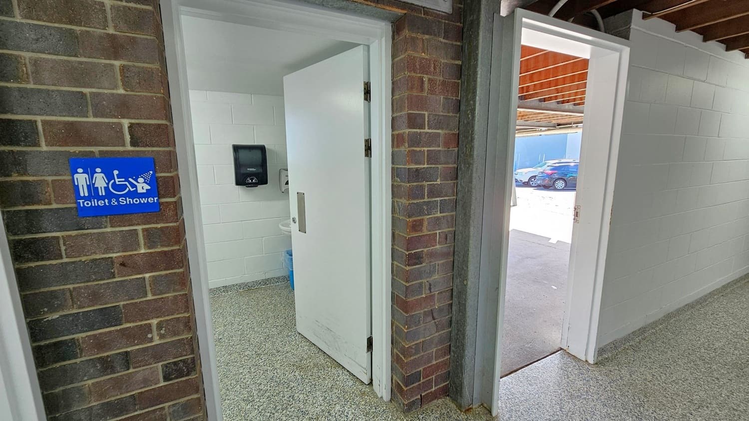A brick wall on the left features a blue sign indicating a toilet and shower, including accessibility symbols. A white door is partially open, revealing a white-tiled interior with a paper towel dispenser. The floor is speckled grey. On the right, another open door leads outside, where several cars are parked. The ceiling has exposed wooden beams.