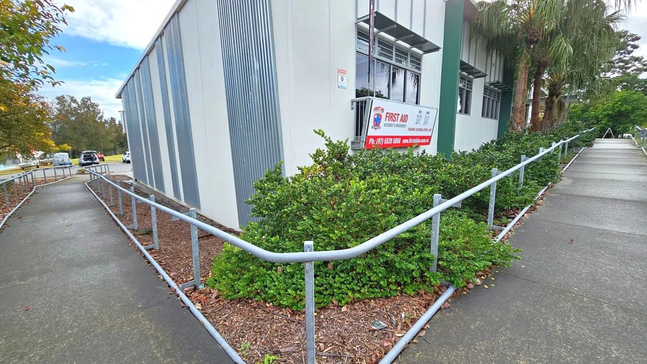 A building with a corrugated metal exterior on the left, featuring a sign for first aid services. A concrete path with metal railings runs alongside the building, bordered by green shrubs. In the background, there are trees and a few parked vehicles. The path leads to a more open area with additional greenery and trees.
