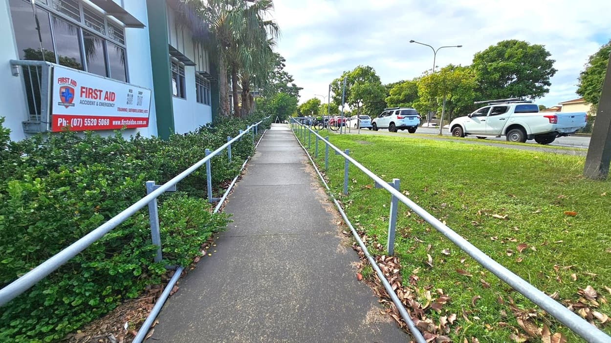 A concrete footpath runs through the centre, bordered by metal railings on both sides. On the left, there's a building with a sign for "First Aid Accident & Emergency" and some green shrubs. On the right, a grassy area with scattered leaves and a few parked white vehicles along the road. Trees line the background, and the sky is overcast.