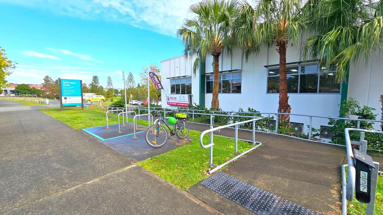 A bicycle with a green helmet is parked at a bike rack on a paved area. To the right, a white building with large windows is partially visible, with palm trees and shrubs in front. A sign for "Sports House Varsity" is on the left, situated on a grassy patch next to a paved path. Several cars are parked in the background, and there are more trees and greenery further back. The ground is a mix of grass and pavement.