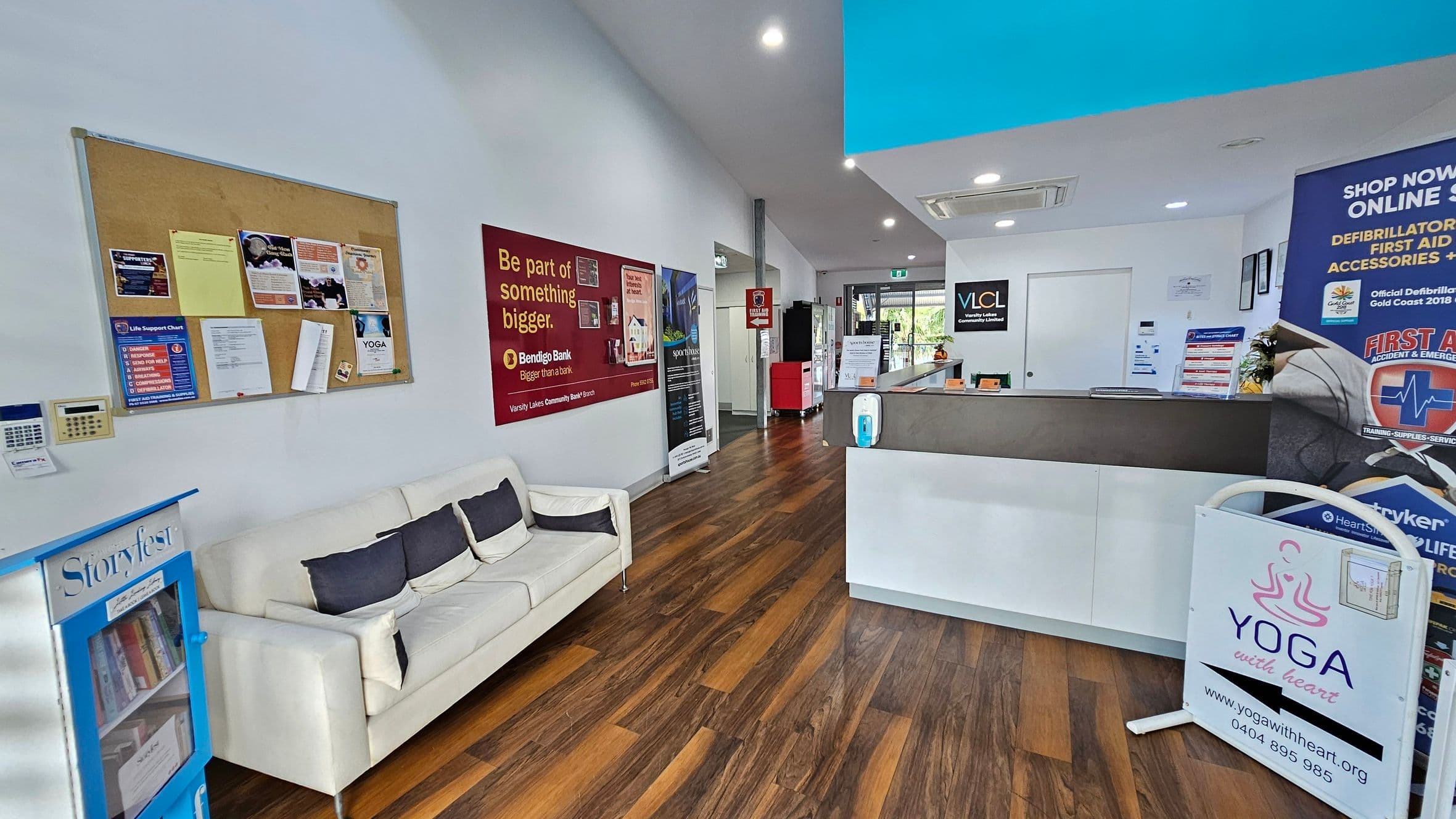 A reception area with wooden flooring and a white ceiling. On the left, a white sofa with dark cushions is placed beneath a bulletin board filled with various notices. A small bookshelf labelled "Storyfest" is nearby. On the right, a reception desk with promotional materials and a sign for "Yoga with Heart" is visible. The back wall features a door and a window, with more posters and a red vending machine. Bright lighting illuminates the space.