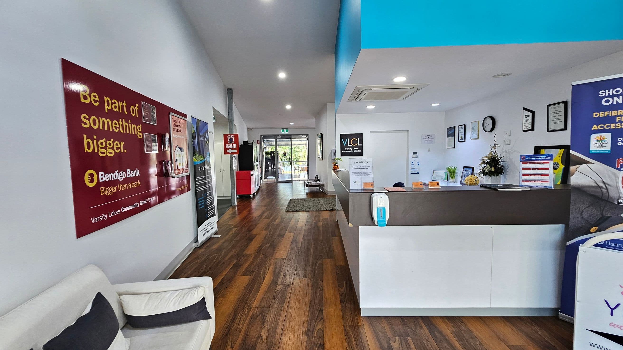 A reception area with a wooden floor and white walls. On the left, a red Bendigo Bank poster is displayed above a white sofa with black and white cushions. The right side features a reception desk with a hand sanitiser dispenser and various informational signs. Behind the desk, framed certificates and a clock are mounted on the wall. The background shows a glass door leading outside, with a small green mat on the floor. The ceiling has recessed lighting.