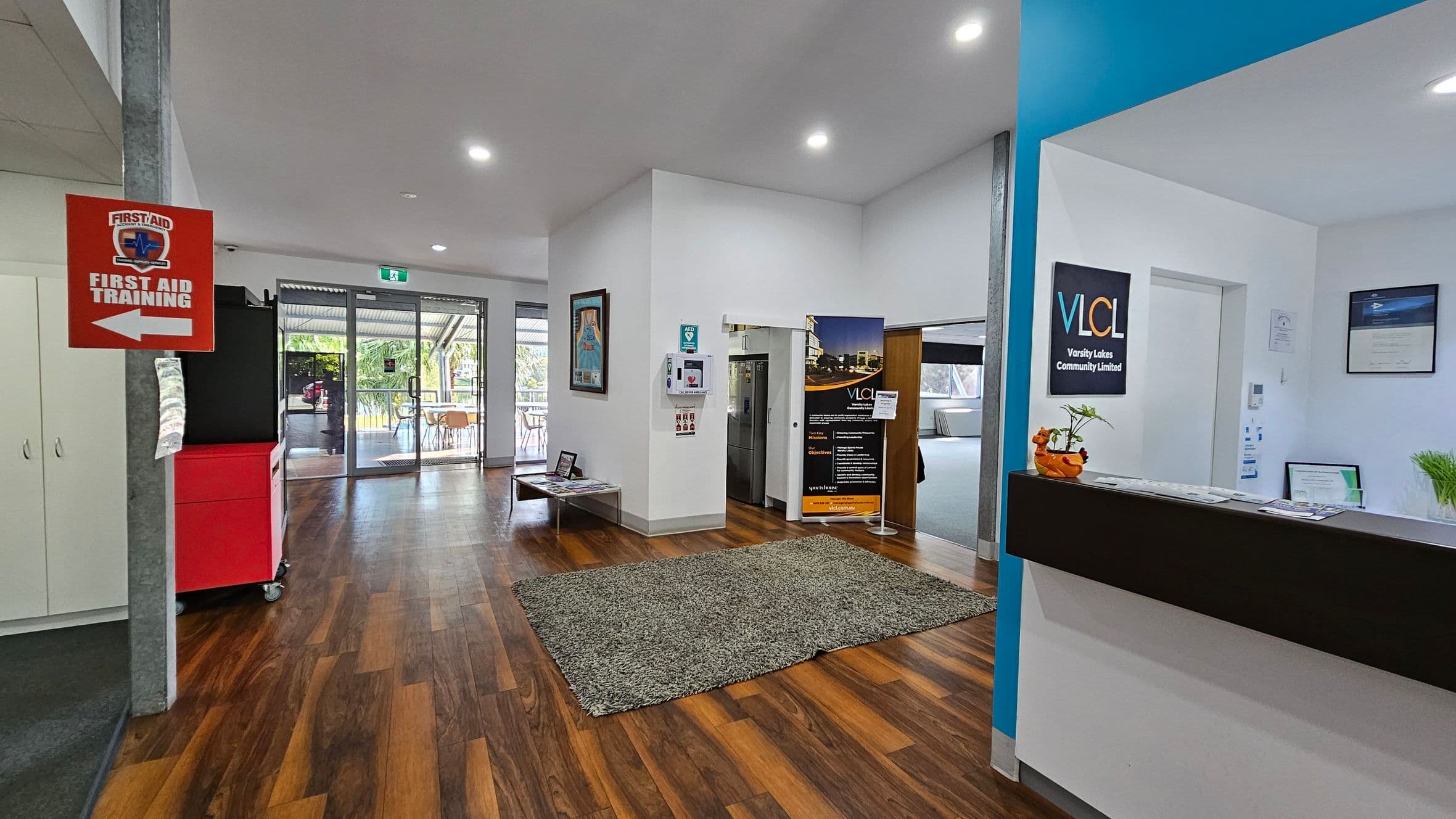 A reception area with a wooden floor and white ceiling. On the left, a red sign indicates "First Aid Training" with an arrow. A red cabinet and white cupboards are nearby. In the centre, a green rug lies on the floor, with a table and posters on the wall. Glass doors lead outside. On the right, a reception desk displays a sign for Varsity Lakes Community Limited, with plants and documents on top. A blue wall section adds contrast.