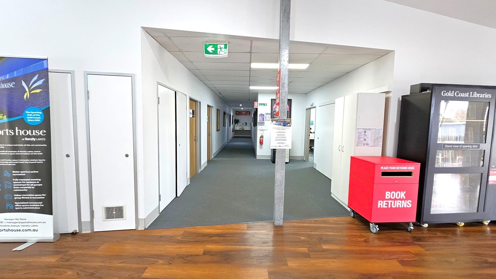A hallway with a wooden floor leading to a carpeted area. On the left, there's a large banner with text and graphics. Several doors line the left wall. On the right, there's a red book return box on wheels and a black library vending machine labelled "Gold Coast Libraries." The ceiling has fluorescent lights and an exit sign. A fire extinguisher is mounted on the wall in the distance.