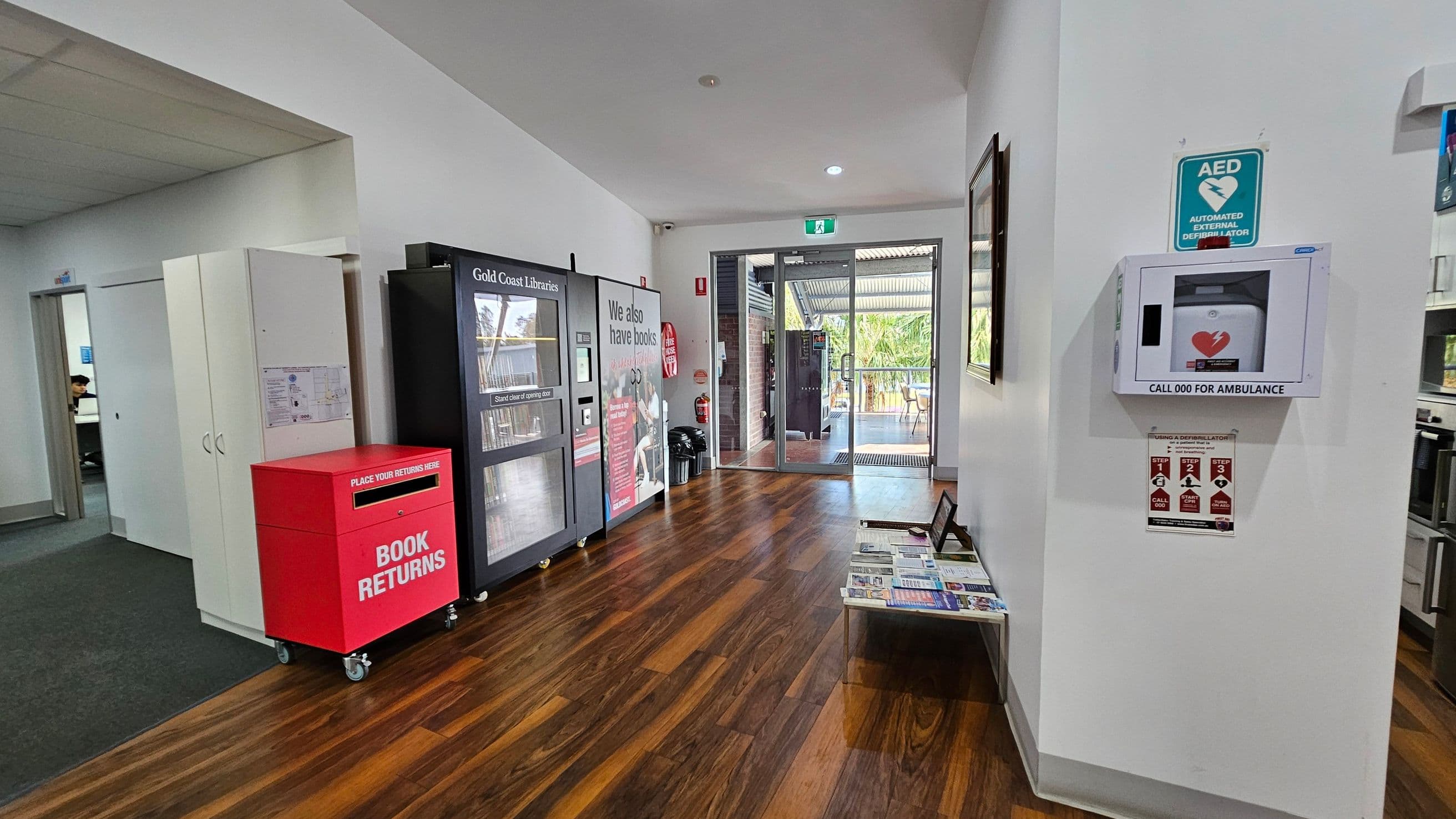 A library entrance area with a wooden floor. On the left, there's a red book return bin and a vending machine for library items. A doorway leads to another room. On the right, a wall-mounted automated external defibrillator (AED) is visible with instructions below. In the centre, glass doors open to an outdoor area with tables and chairs. A small table with brochures is against the right wall. The ceiling is white with recessed lighting.