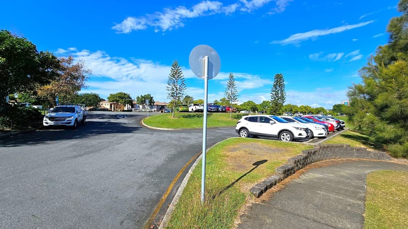A curved road with a white vehicle parked on the left side. In the centre, a metal pole with a circular sign. On the right, a row of parked cars in various colours, including white and red, on a grassy area. Trees and shrubs are scattered throughout, with a few tall, narrow trees in the background. A paved footpath runs alongside the grass, bordered by a low brick wall.