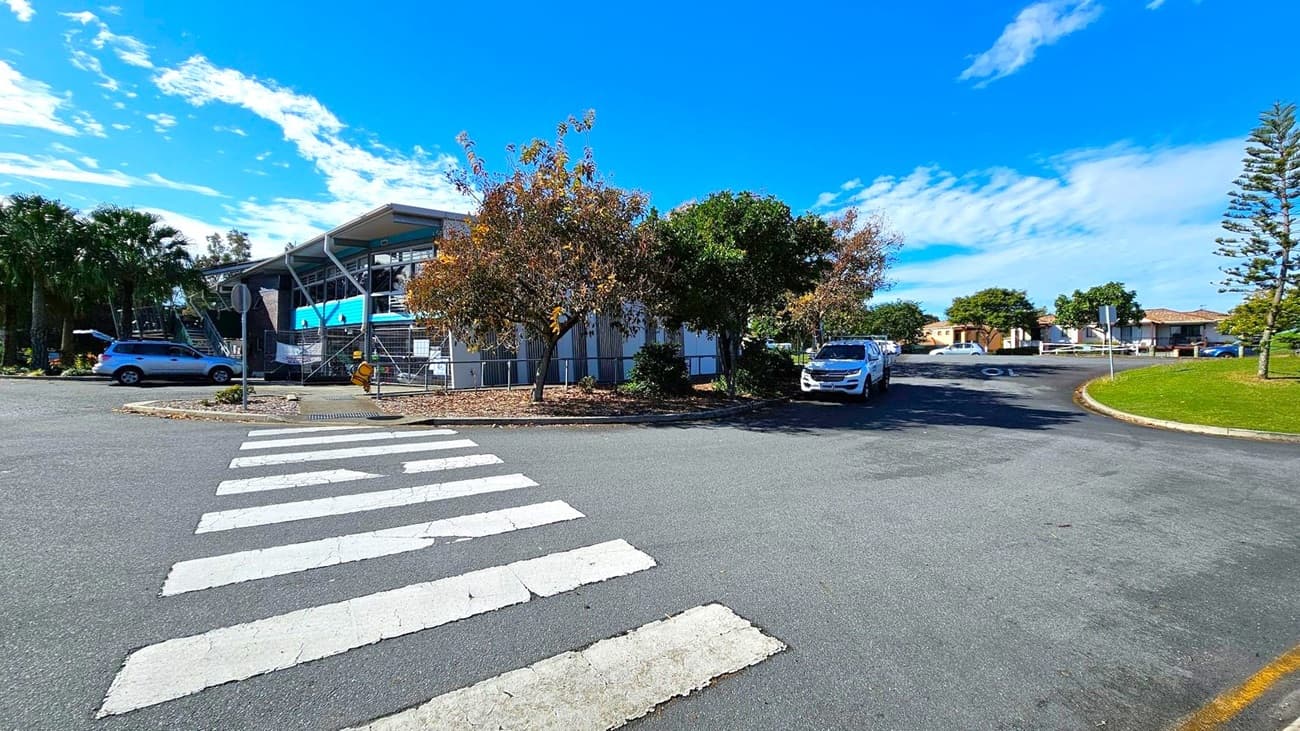 A zebra crossing leads to a building with a modern design on the left, featuring large windows and a slanted roof. Several trees with autumn foliage are in front of the building. A silver car is parked nearby. On the right, a white vehicle is parked along a curved road, with a grassy area and more trees. In the background, there are residential houses with tiled roofs. The sky is clear with a few clouds.