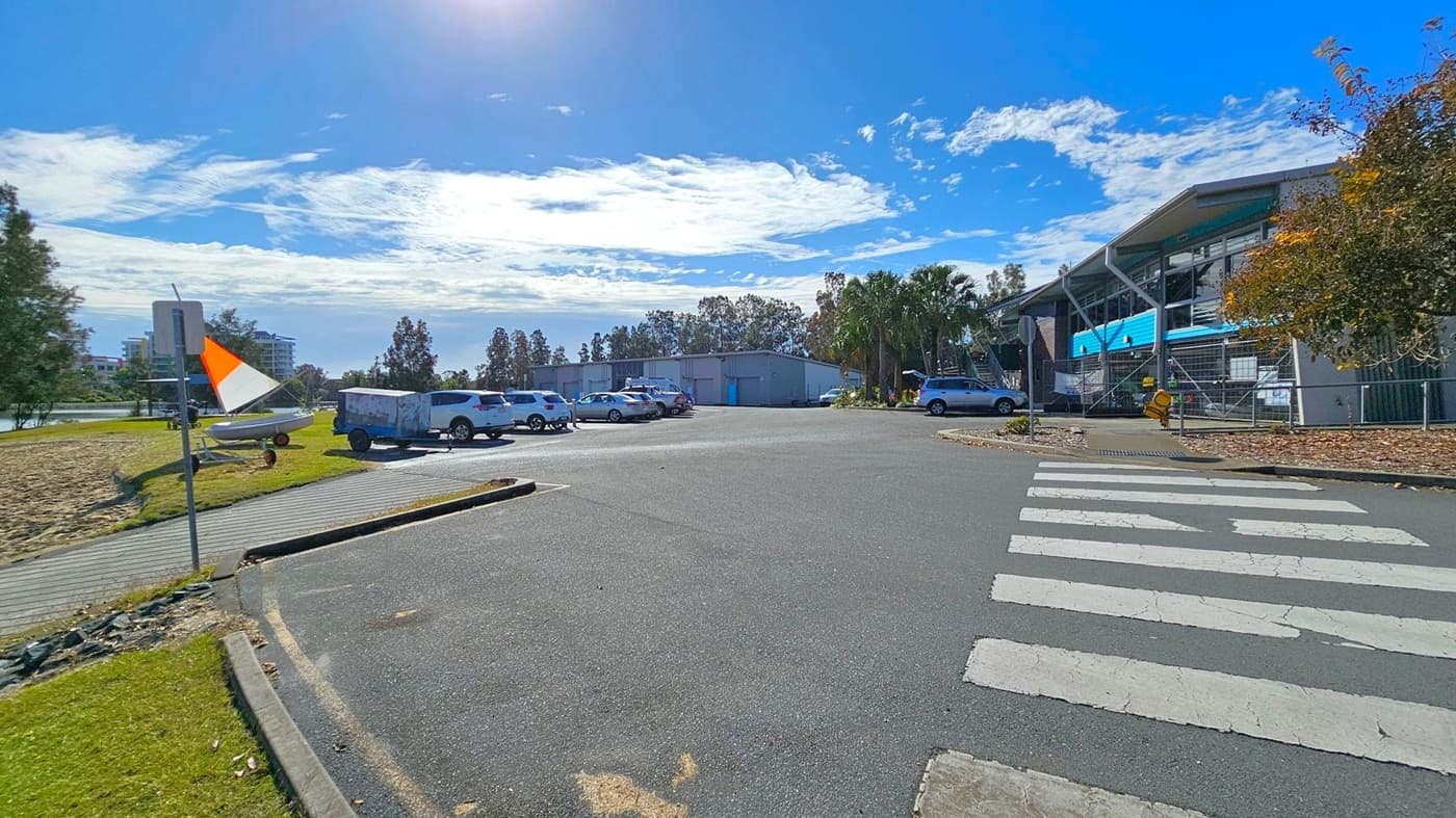 A car park with several vehicles is in the centre, surrounded by trees and a building on the right with a blue facade. A pedestrian crossing is visible in the foreground. On the left, a grassy area with a small boat and a trailer is near a path. A sign with an orange and white flag is also on the left. The background features more trees and a few buildings.