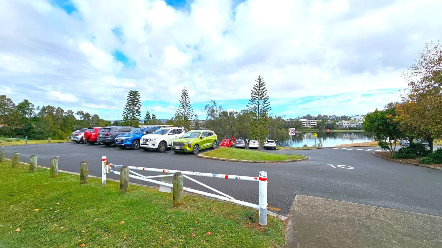 A car park with several vehicles, including a bright green car, is on the left. The area is bordered by wooden posts and a white metal gate with red markings. In the background, there are tall trees and a body of water with buildings visible on the far side. The road in the foreground has a speed limit sign marked "10". The scene is surrounded by greenery, with trees on the right.