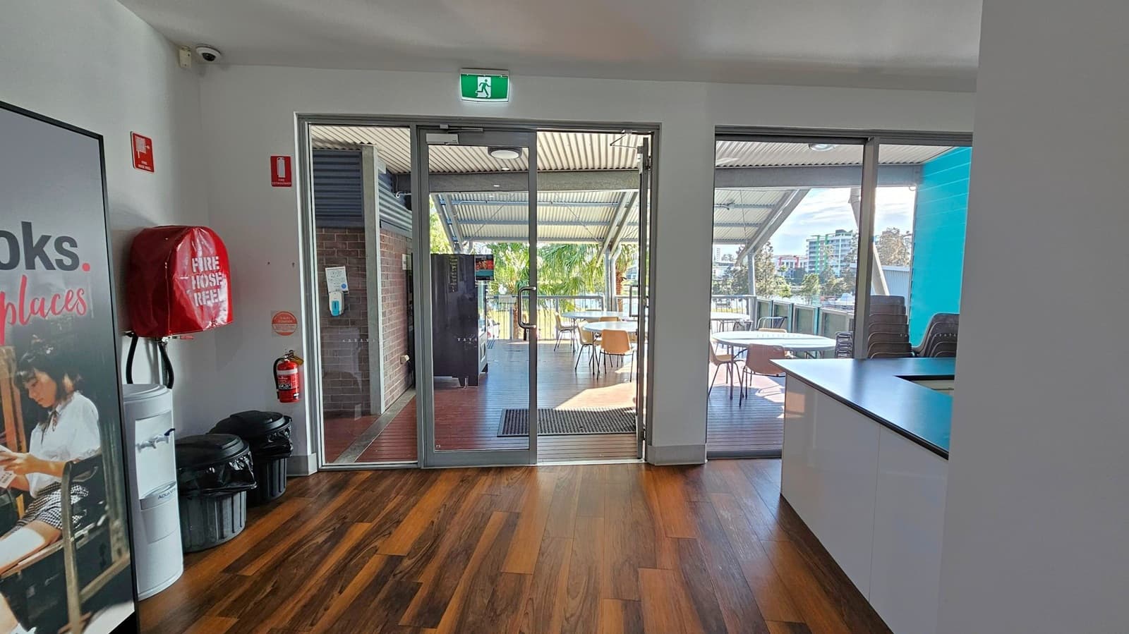 A room with wooden flooring and a white ceiling. On the left, there's a poster, a fire hose reel, a water cooler, and two bins. In the centre, glass doors lead to an outdoor area with tables and chairs under a metal roof. A fire extinguisher is mounted on the wall near the door. On the right, there's a countertop with a glossy finish. Outside, trees and buildings are visible in the background.