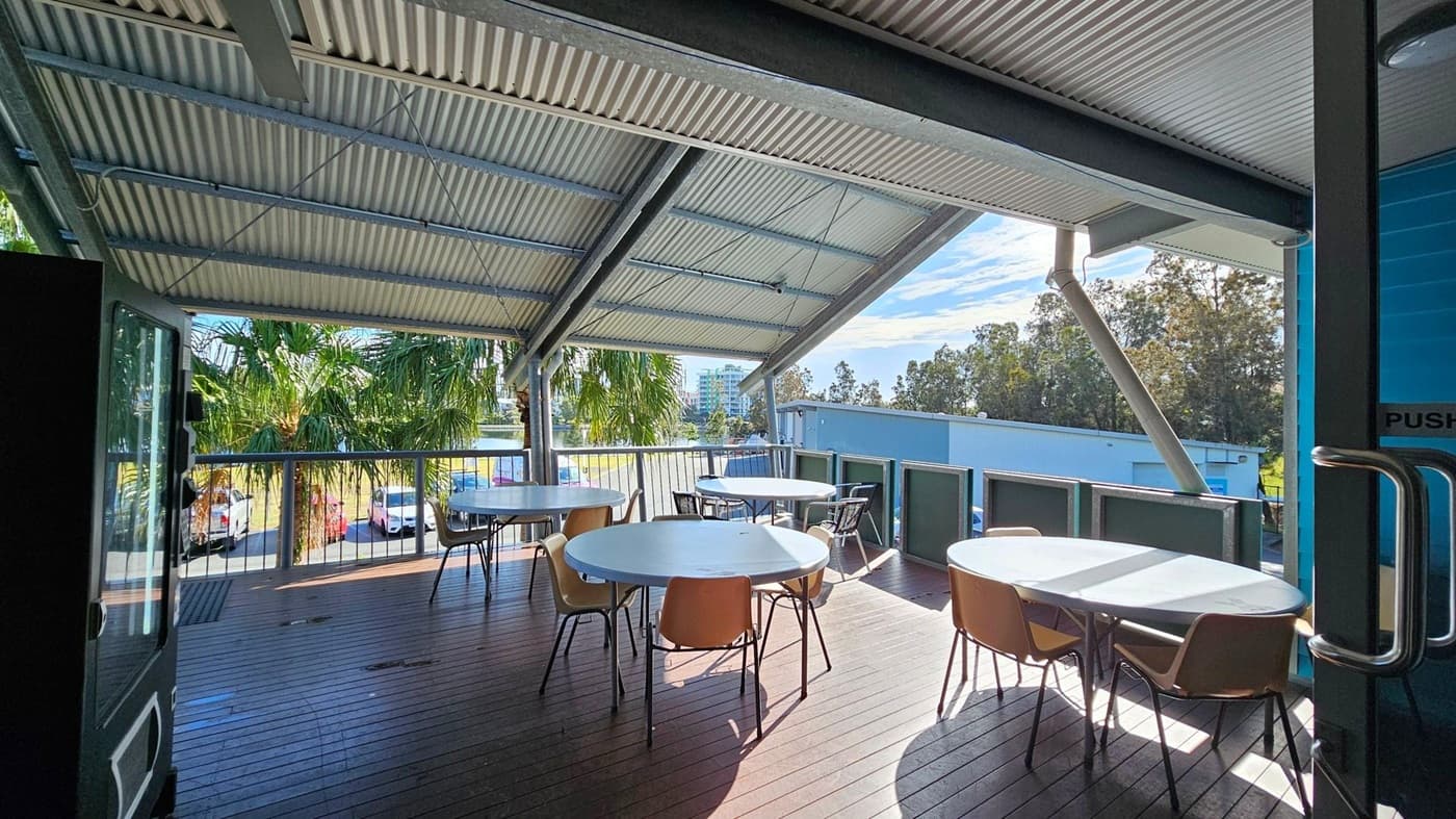 A covered outdoor deck with a corrugated metal roof and wooden flooring. Several round tables with chairs are arranged on the deck. The area is surrounded by a railing, and palm trees are visible in the background. Beyond the trees, there are parked cars and a glimpse of buildings. A glass door with a metal handle is partially open on the right side.