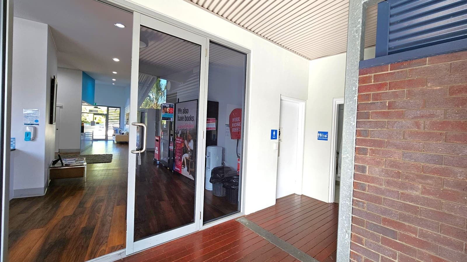 On the left, there is a glass door with a metal handle leading into a room with wooden flooring. Inside, a wall-mounted screen and a hand sanitiser dispenser are visible. In the middle, a sign reads "We also have books" next to a fire hose reel. On the right, there are two doors with blue signs indicating restrooms. The floor outside the doors is wooden, and the ceiling has a light-coloured, ribbed design. A brick wall is partially visible on the far right.