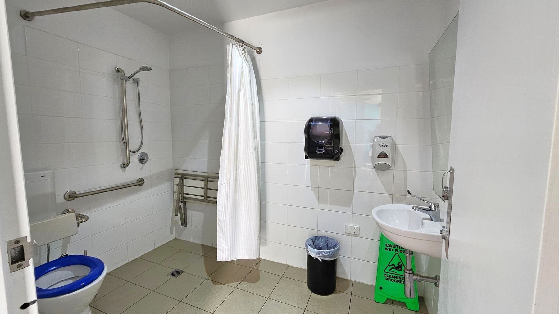 Accessible bathroom with white tiled walls and a light green tiled floor. On the left, a toilet with a blue seat and support bars. A shower area with a handheld showerhead and a white curtain. In the centre, a foldable shower seat. On the right, a sink with a soap dispenser and a paper towel dispenser above a bin. A green "Caution: Wet Floor" sign is beneath the sink. The ceiling is plain white.