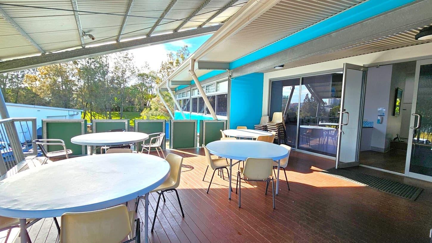 A covered outdoor seating area with several round tables and chairs on a wooden deck. On the right, there are glass doors leading inside a building with a blue exterior. The ceiling is metal, and the area is partially enclosed with green panels. In the background, there are trees and a grassy area, with a white building visible to the left.