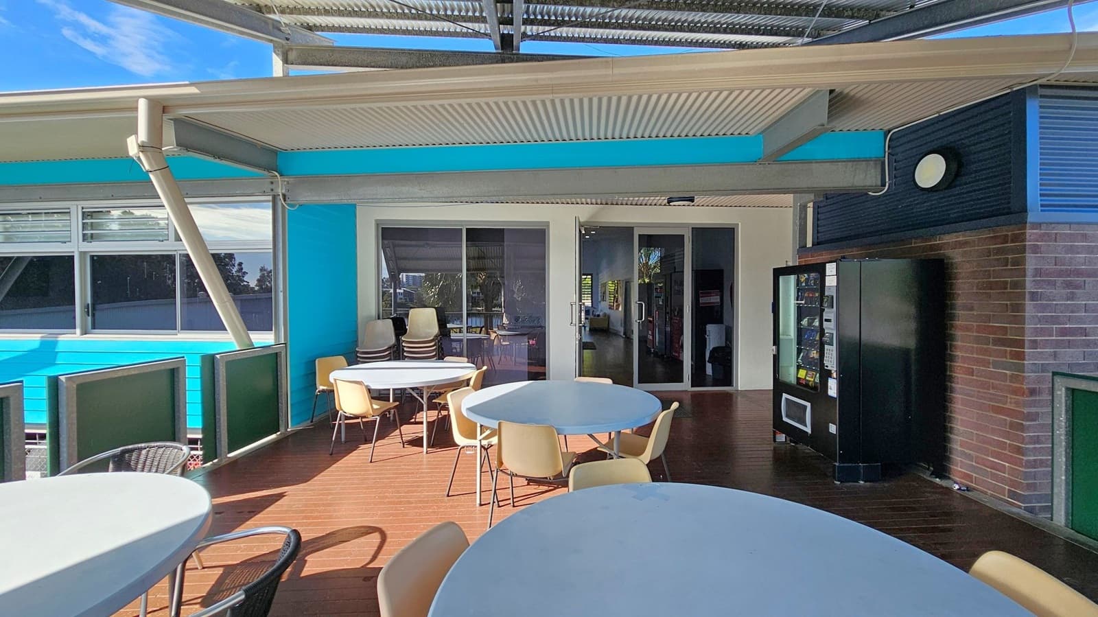 Outdoor seating area with round tables and chairs on a wooden deck. On the right, a black vending machine is positioned against a brick wall. The background features a blue and white building with large windows and a glass door. The ceiling is made of corrugated metal, supported by beams. A stack of chairs is visible near the door.