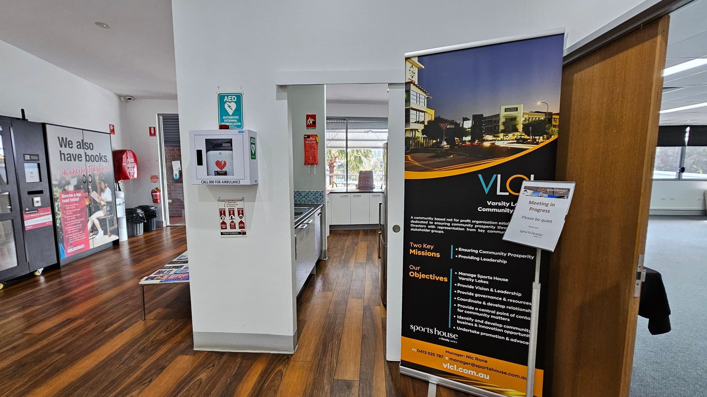 A room with wooden flooring and white walls. On the left, a large poster with text about books and a vending machine. In the centre, an AED mounted on the wall and a small kitchen area with a sink and window. On the right, a large banner with information about Varsity Lakes Community Limited and a sign indicating a meeting in progress. A partially open wooden door leads to a carpeted area.