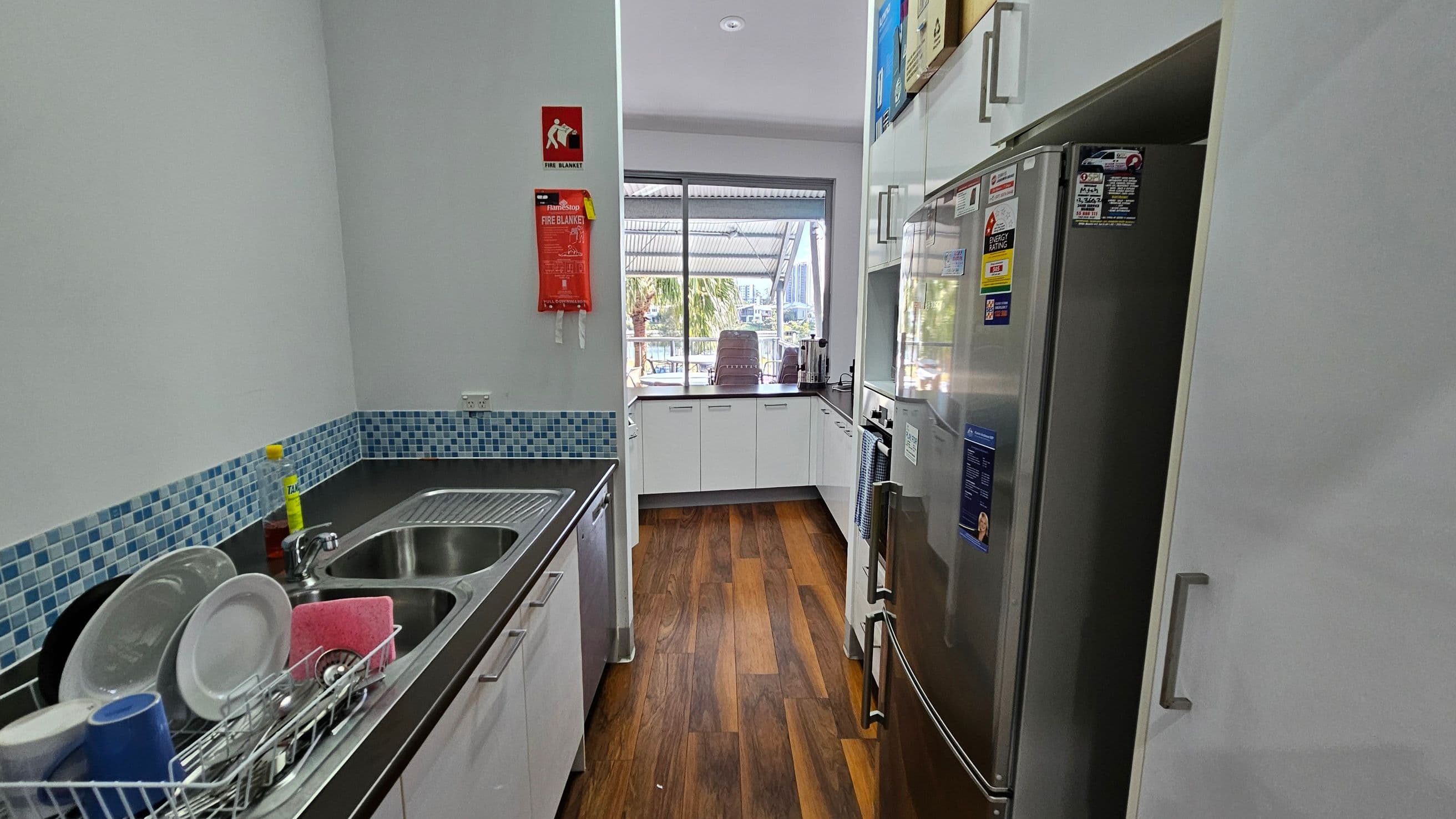 A narrow kitchen with a wooden floor. On the left, there's a countertop with a double sink, a dish rack with plates and cups, and a bottle of dish soap. Above the sink is a blue mosaic tile backsplash. On the right, a stainless steel refrigerator with various stickers. In the centre, a window with a view of outdoor seating and palm trees. A fire blanket is mounted on the wall near the window. White cabinets line the walls, and the ceiling has recessed lighting.