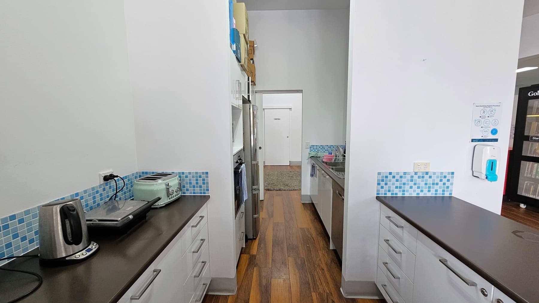 A narrow kitchen with wooden flooring and white walls. On the left, a black countertop holds a stainless steel kettle, a sandwich press, and a mint green toaster. Above are blue mosaic tiles. On the right, a similar countertop with a sink and dish rack. A hand sanitiser dispenser and a poster are mounted on the wall. In the background, a doorway leads to a room with a green rug. Shelves and a stainless steel fridge are visible along the left wall.