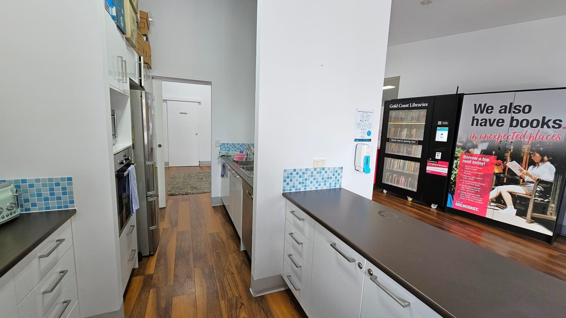 A modern kitchen area with white cabinets and black countertops. On the left, there is a microwave and a fridge, with a blue and white checkered towel hanging from the oven handle. The floor is wooden. In the background, a green rug leads to a white door. On the right, a vending machine with books and a large poster advertising library services. Blue mosaic tiles are used as a backsplash above the sink and counter. A hand sanitiser dispenser is mounted on the wall.