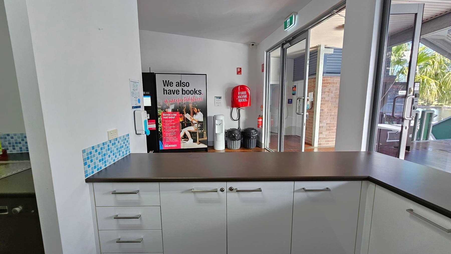 A room with a countertop in the foreground, featuring white cabinets with silver handles. On the left, there's a wall with a blue-tiled backsplash and a sink. In the centre, a poster with text and a fire hose reel are mounted on the wall. To the right, glass doors lead outside, showing a view of a brick wall and palm trees. The ceiling has a white finish, and the floor is wooden. An exit sign is above the doors.