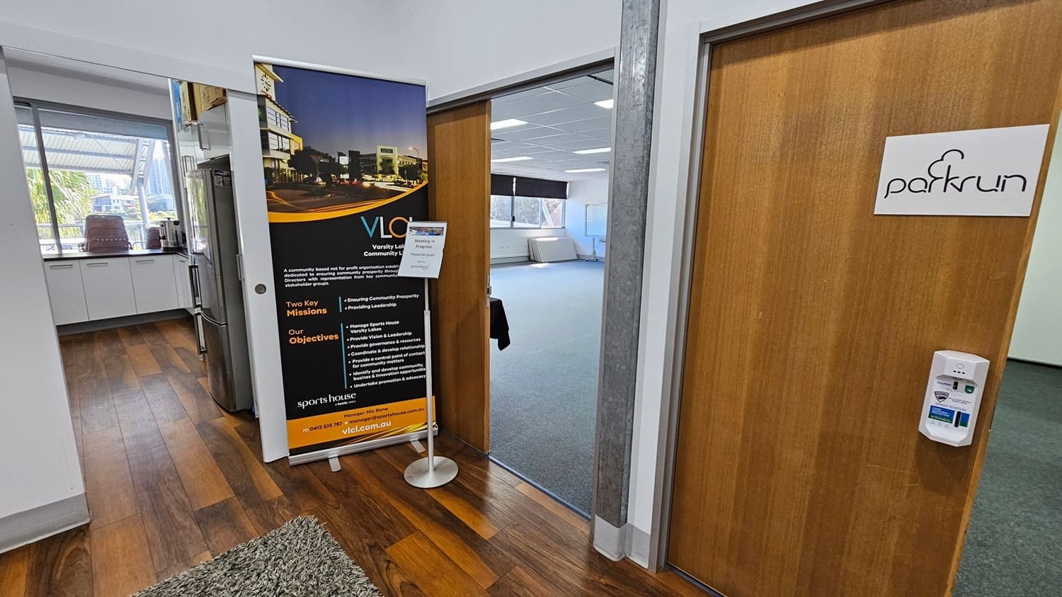 A hallway with wooden flooring and a green rug. On the left, there's a kitchen area with white cabinets, a refrigerator, and a window showing an outdoor view with trees and buildings. In the centre, a large vertical banner with text and images stands next to an open door leading to a room with grey carpet and a window. On the right, a wooden door with a "parkrun" sign and a wall-mounted device is visible.