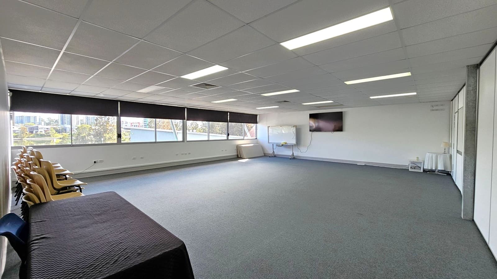 A spacious room with a grey carpeted floor and a grid-patterned ceiling with fluorescent lights. On the left, there are stacked wooden chairs and a table covered with a black cloth. Large windows with black blinds line the left wall, offering a view of buildings and trees outside. On the right, a whiteboard and a wall-mounted TV are visible. A small round table with a white cloth is in the corner. The room is mostly empty, providing an open area.