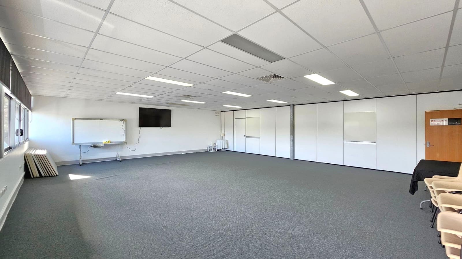 A spacious room with a grey carpeted floor and a white tiled ceiling featuring fluorescent lights. On the left, there are large windows with blinds, a whiteboard, and a wall-mounted TV. The right side has a wooden door and a stack of chairs. The back wall is lined with white partition panels, and a small table with items is visible.