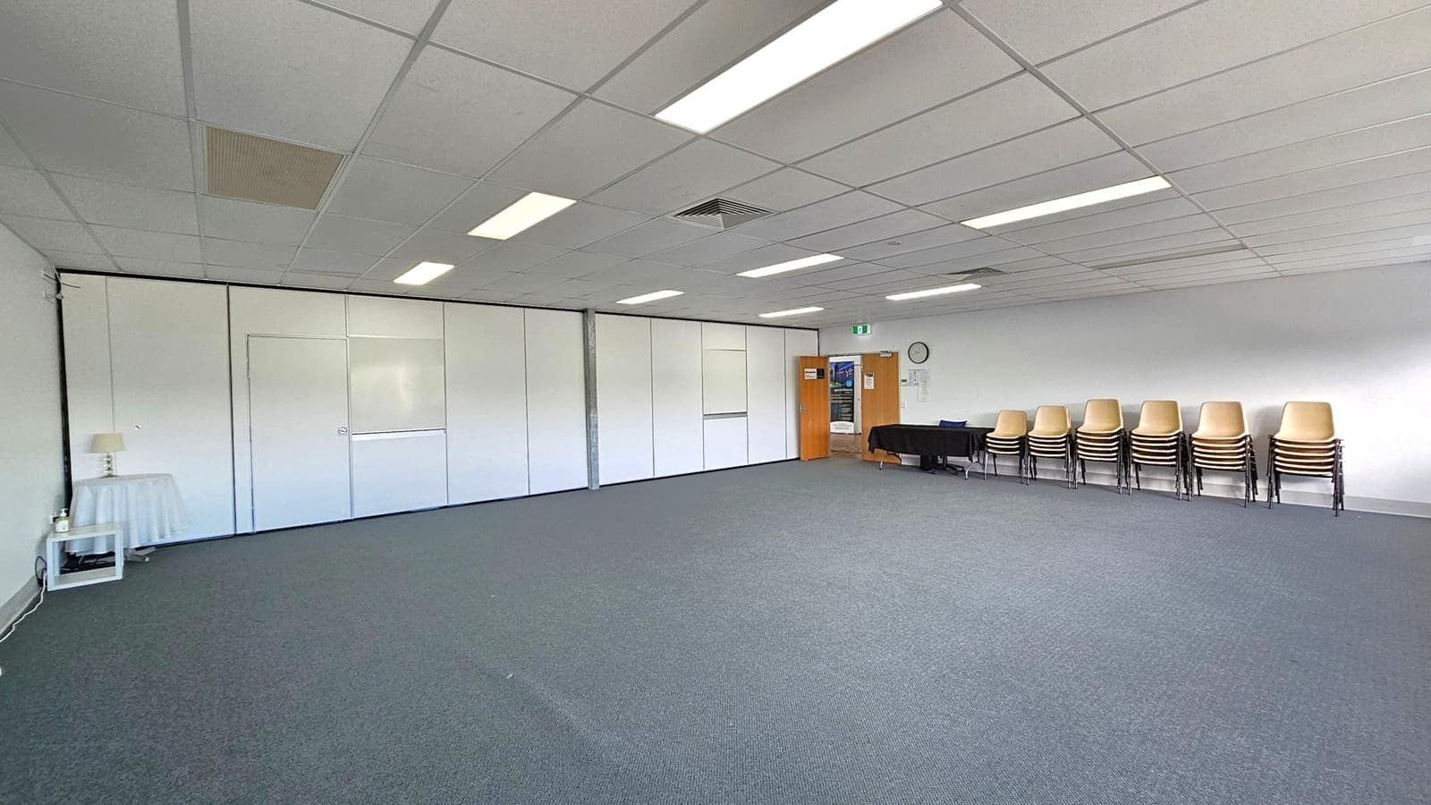 A spacious room with a grey carpeted floor and a white tiled ceiling featuring fluorescent lights. On the right, there are stacks of chairs against the wall, next to a black table. A clock is mounted on the wall above. In the background, a wooden door with a poster leads to another area. On the left, a small table with a lamp and a bottle is placed against a partitioned wall.