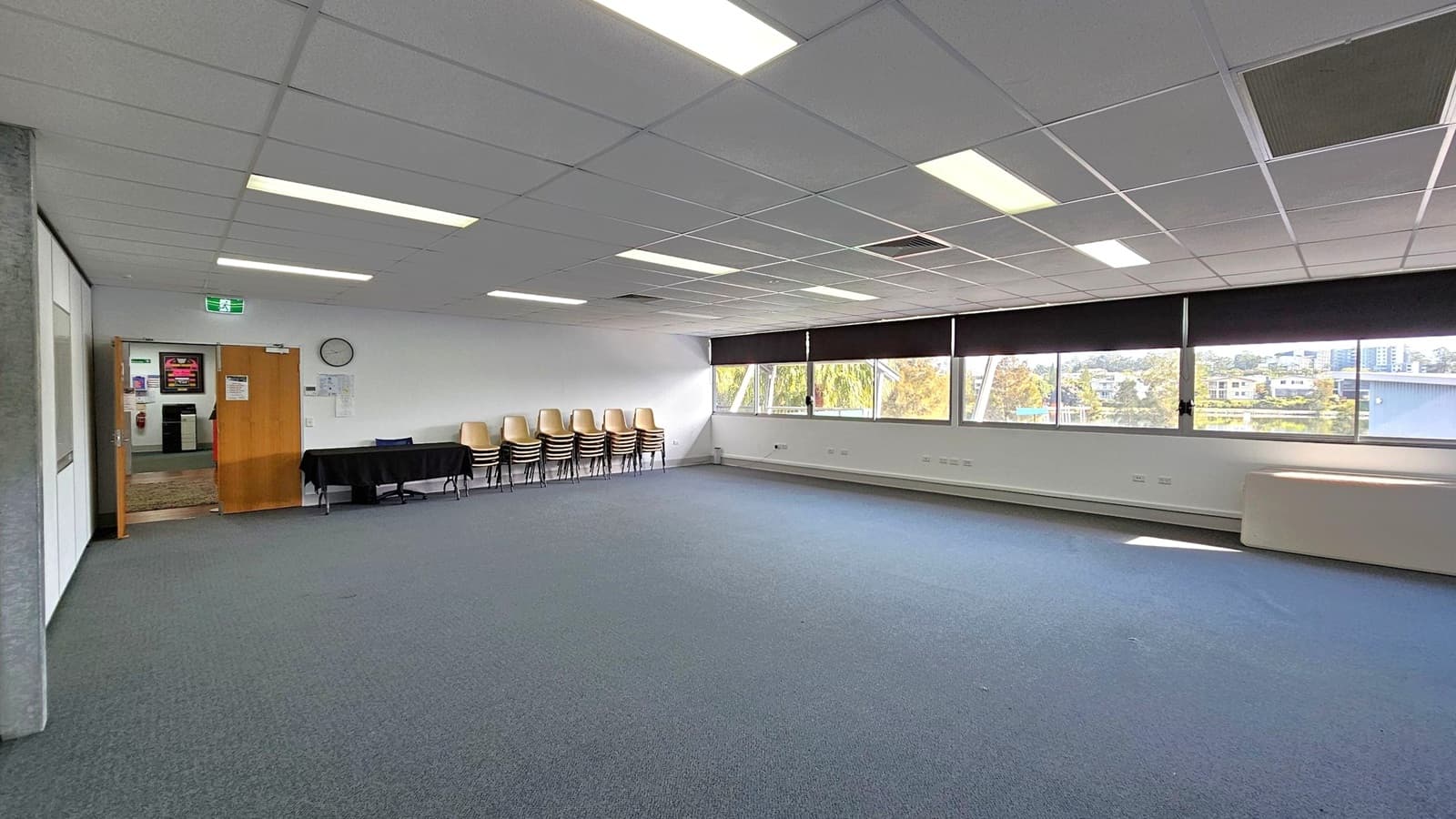 A spacious room with a grey and blue carpeted floor. On the left, there is an open wooden door leading to another area, with a clock and some notices on the wall. A black table with stacked chairs is nearby. The right side features large windows with black blinds, offering a view of trees and buildings outside. The ceiling is fitted with fluorescent lights and ceiling tiles.