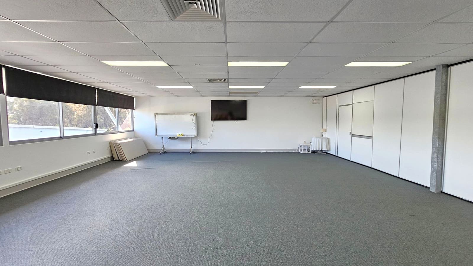 A spacious room with a grey carpeted floor and a white tiled ceiling featuring fluorescent lights. On the left, there are large windows with black blinds, and several folded tables are stacked against the wall. In the centre, a whiteboard and a wall-mounted TV are visible. On the right, there are white partition walls with a door. A small table with a cloth and some items is near the corner.