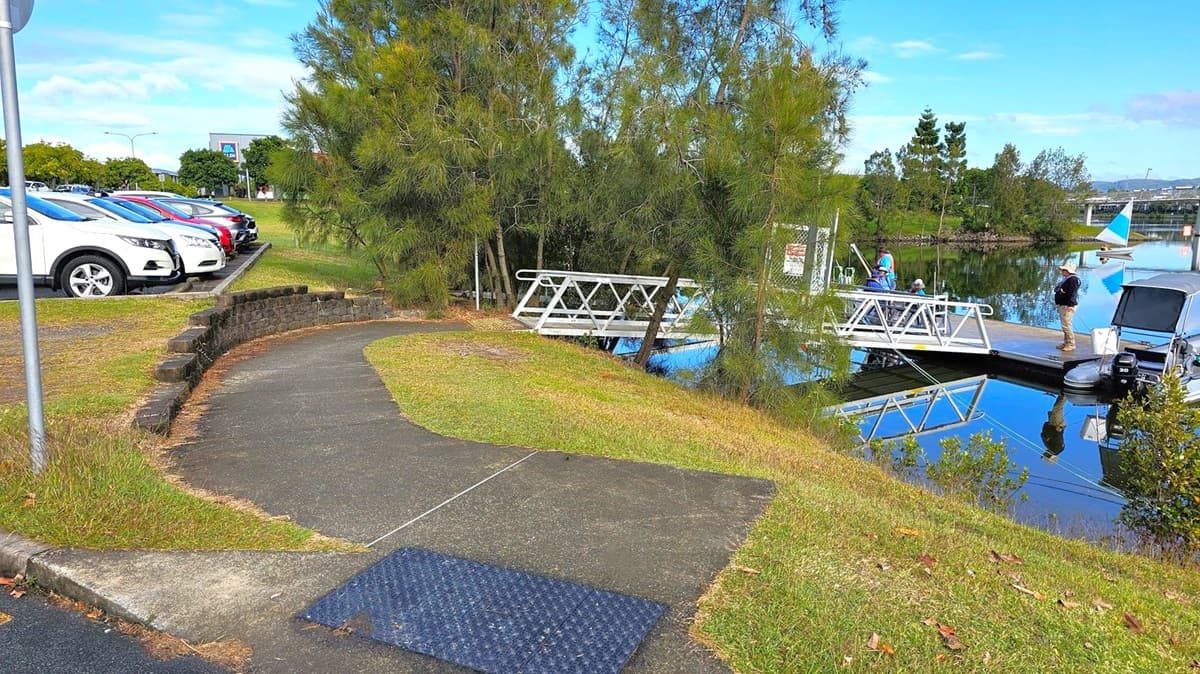 A curved path leads towards a small dock on the right, where a few people are standing near a boat. The dock is surrounded by calm water reflecting the scene. On the left, a row of parked cars is visible, with a grassy area and a low stone wall separating the path from the parking lot. Trees are scattered throughout the area, providing some shade. In the background, a small sailboat is on the water, and a bridge is visible in the distance.