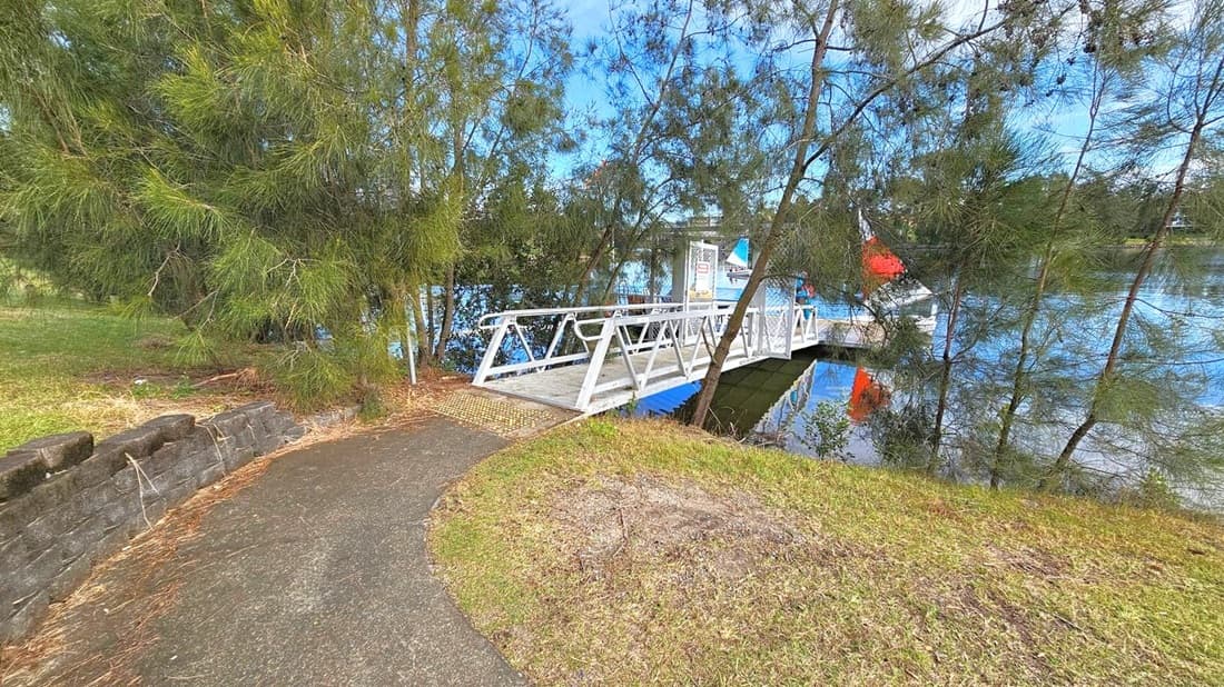 A narrow path leads to a white metal bridge over a body of water. On the left, dense green trees and a low stone wall line the path. The bridge extends to a small dock where a person stands near a sailboat with a red and black sail. The water reflects the trees and the boat. The ground is grassy with patches of dirt.