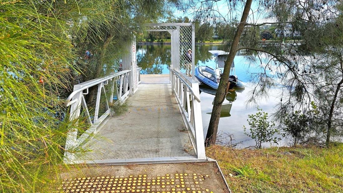 A metal walkway with railings extends towards a small dock over a calm body of water. On the left, dense green foliage partially obscures the view. On the right, a tree with thin branches leans over the water, where a small boat is moored. The background features a reflective water surface with trees lining the opposite bank. The ground near the walkway is grassy with a tactile paving surface at the entrance.