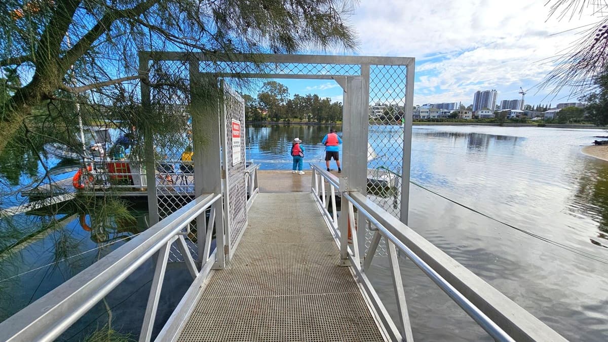 A metal walkway with a mesh gate leads to a dock over a body of water. Two people in life jackets stand at the end of the dock, facing the water. On the left, a tree with dense branches partially obscures the view. In the background, across the water, there are buildings and a crane, indicating an urban area. The water reflects the surroundings, and the sky is partly cloudy.