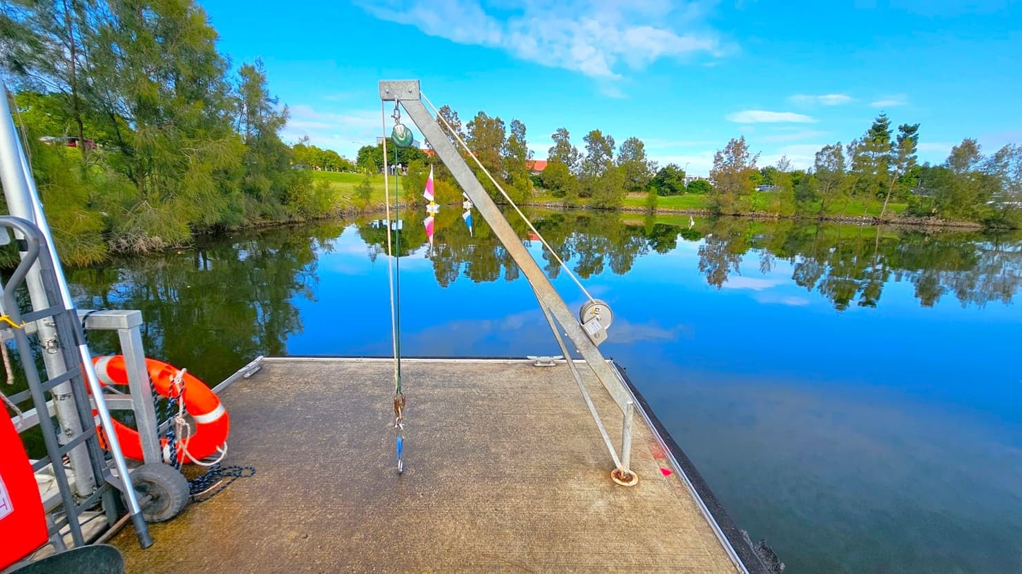 A calm lake with clear reflections of trees and a grassy bank in the background. In the foreground, a concrete platform extends over the water, featuring a metal crane with a pulley system and a hook. To the left, a red and white lifebuoy is attached to a metal structure with wheels. The sky is blue with scattered clouds, and the overall scene is tranquil and still.