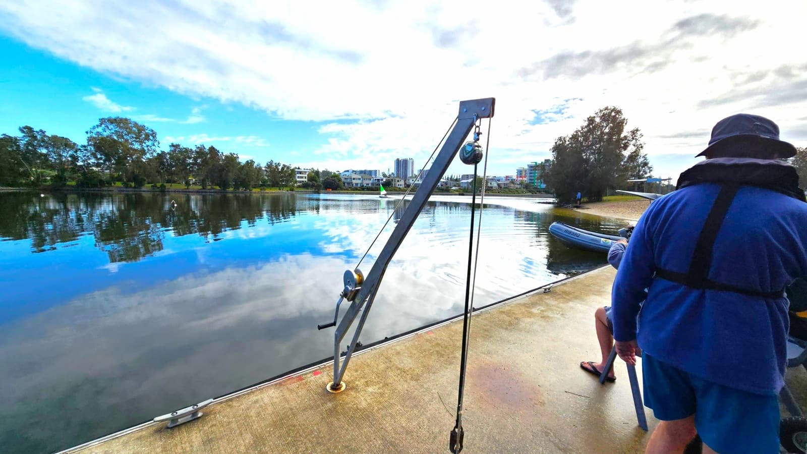 A calm body of water reflects the sky and surrounding trees. On the left, a metal crane with a pulley system is mounted on a concrete surface. In the background, modern buildings are visible across the water. On the right, a person wearing a blue outfit and hat stands near the edge, with another person seated nearby. A small inflatable boat is partially visible near the shore. Trees line the far bank, and the sky is partly cloudy.