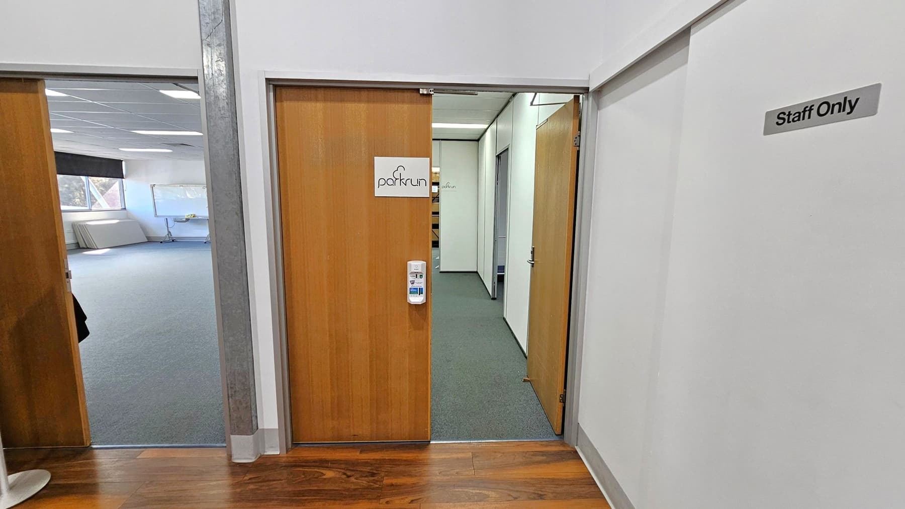 A hallway with two wooden doors. The door on the right has a sign reading "parkrun" and a hand sanitiser dispenser mounted on it. The wall next to it has a "Staff Only" sign. The door on the left opens into a room with grey carpet, a large window, and a whiteboard. The floor in the hallway is wooden, and the ceiling has recessed lighting.