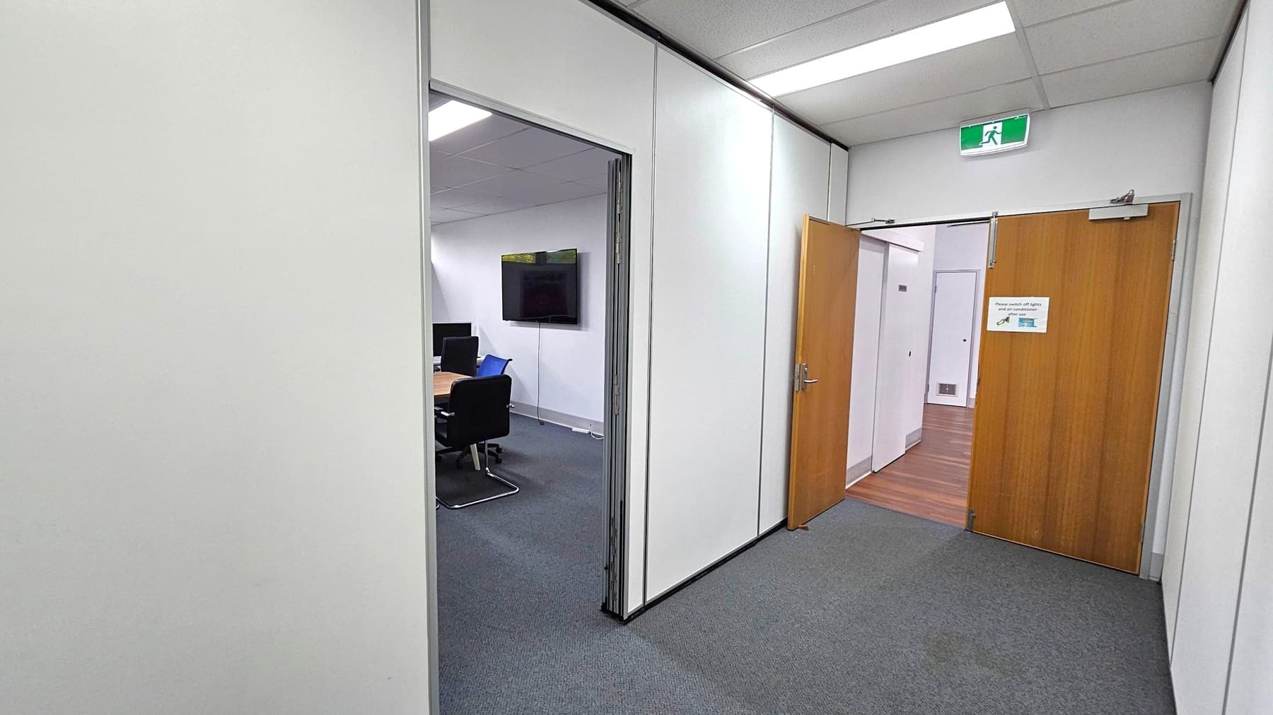 A corridor with grey carpeted flooring and white walls. On the right, a wooden door is partially open, revealing a room with wooden flooring. Above the door, an emergency exit sign is visible. On the left, an open doorway leads to a meeting room with a table, chairs, and a wall-mounted screen. The ceiling has fluorescent lighting.